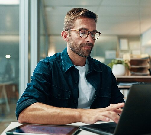 Ein Mann mit Brille sitzt konzentriert an einem Schreibtisch und tippt auf seinem Laptop in einem Büroumfeld.  Das Bild zeigt einen Mann mittleren Alters mit kurzem, leicht grau meliertem Haar und einem gepflegten Bart. Er trägt eine Brille mit gemustertem Rahmen, ein weißes T-Shirt und darüber ein dunkelblaues Jeanshemd. Seine Haltung deutet auf Konzentration hin, während er auf einem schwarzen Laptop tippt, der vor ihm auf dem Schreibtisch steht. Links neben dem Laptop befindet sich ein Tablet.  Der Hintergrund ist leicht unscharf, aber man erkennt, dass er sich in einem Büro befindet. Im Hintergrund sind Büromöbel, Aktenordner und eine Zimmerpflanze zu sehen. Glaswände deuten darauf hin, dass es sich um ein modernes Büro handelt.  Die Atmosphäre des Bildes ist professionell und fokussiert. Die Farbgebung ist gedämpft und die Beleuchtung neutral, was zur Ernsthaftigkeit der Szene beiträgt. Die Stimmung ist ruhig und konzentriert, was die Arbeitsatmosphäre widerspiegelt.