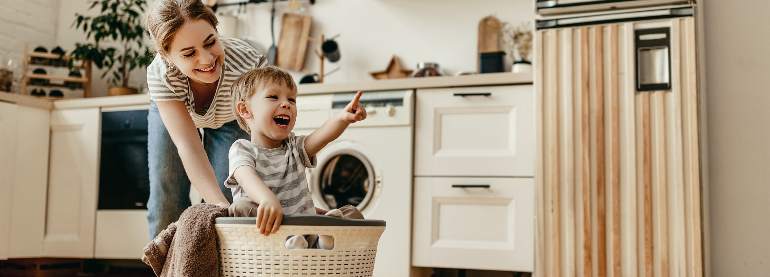 Ein Kind sitzt in einem Wäschekorb und zeigt mit dem Arm nach vorne. Eine erwachsene Person steht dahinter und hält den Korb fest. Die Szene spielt in einer Küche mit weißen Schränken, einem Backofen und einer Waschmaschine. Rechts ist eine Holzverkleidung an der Wand zu sehen.